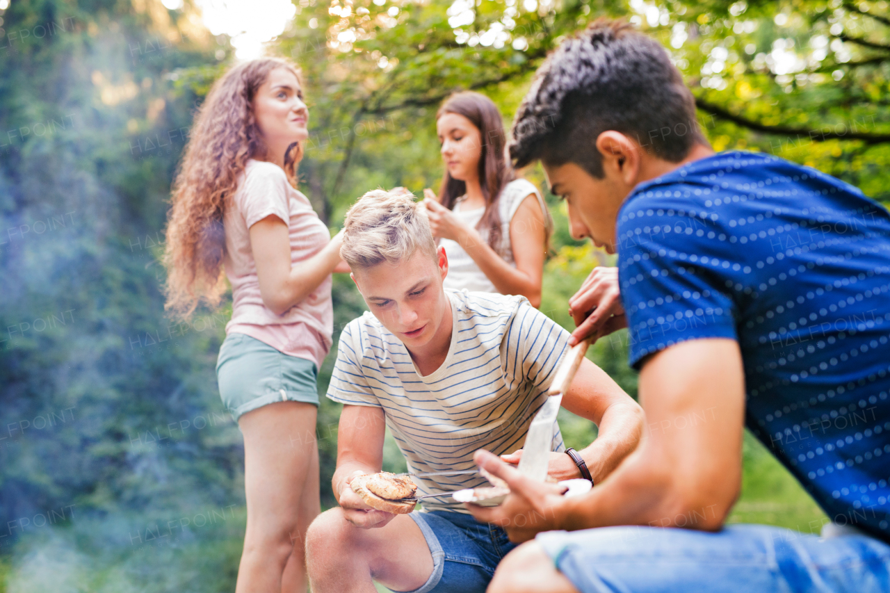 Beautiful teenagers enjoying camping holiday in forest. Boys cooking meat on barbecue grill.