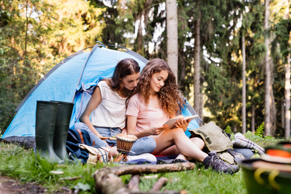 Two teenage girls camping in forest, sitting in front of tent, reading a book. Summer vacation adventure.