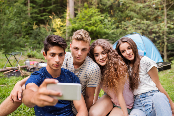 Group of teenagerss camping in forest, sitting in front of tent, taking selfie with smart phone. Summer vacation adventure.