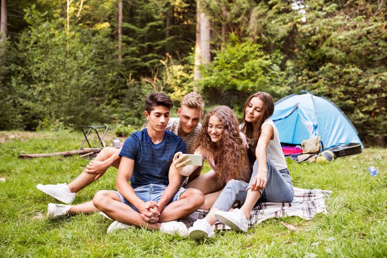 Group of teenagerss camping in forest, sitting in front of tent, taking selfie with smart phone. Summer vacation adventure.