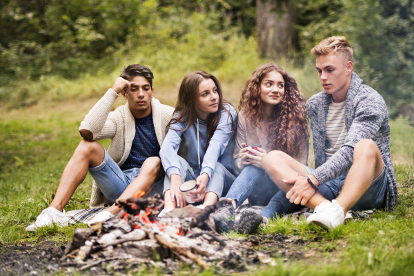 Beautiful teenagers enjoying camping vacations in forest, sitting at bonfire.