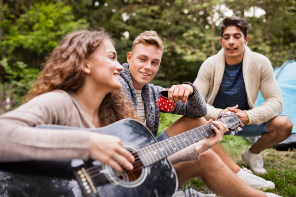 Group of teenagers camping in forest, sitting in front of tent, girl playing guitar.