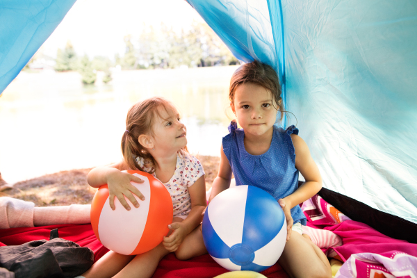 Two beautiful little girls with colorful beach balls sitting in tent, camping by the lake.