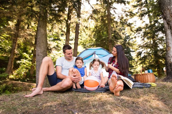 Beautiful young family with cute little daughters camping in forest, sitting in front of tent.