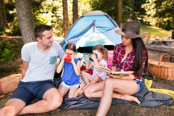 Beautiful young family with cute little daughters camping in forest, sitting in front of tent.