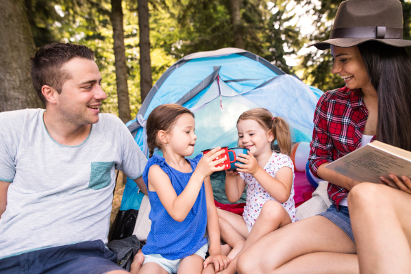 Beautiful young family with cute little daughters camping in forest, sitting in front of tent, girls drinking water.