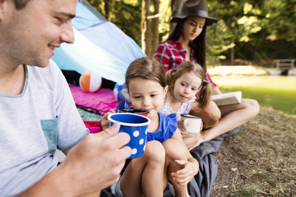 Beautiful young family with cute little daughters camping in forest, sitting in front of tent.