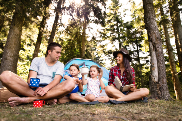 Beautiful young family with cute little daughters camping in forest, sitting in front of tent.