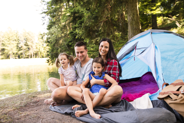 Beautiful young family with cute little daughters camping in forest, sitting in front of tent.