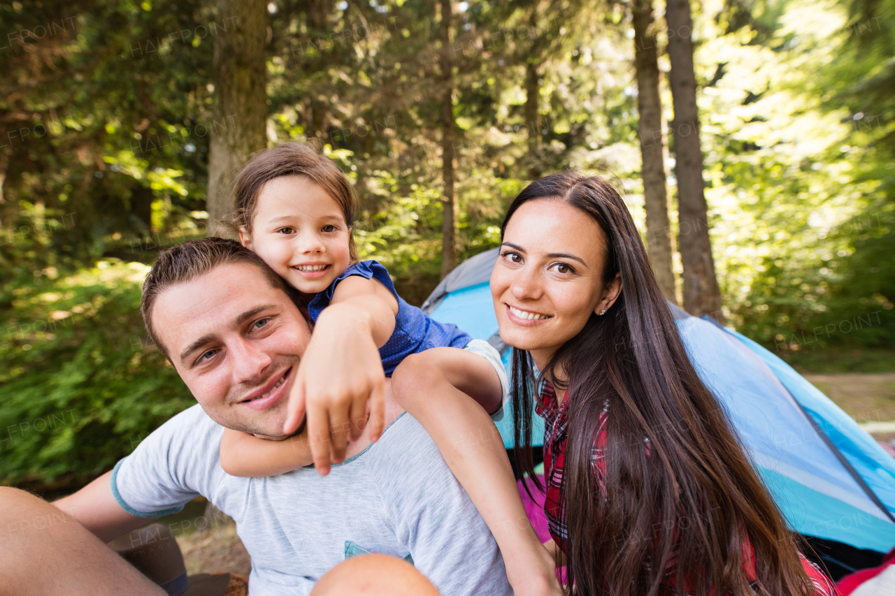 Beautiful young family with cute little daughter camping in forest, sitting in front of tent.