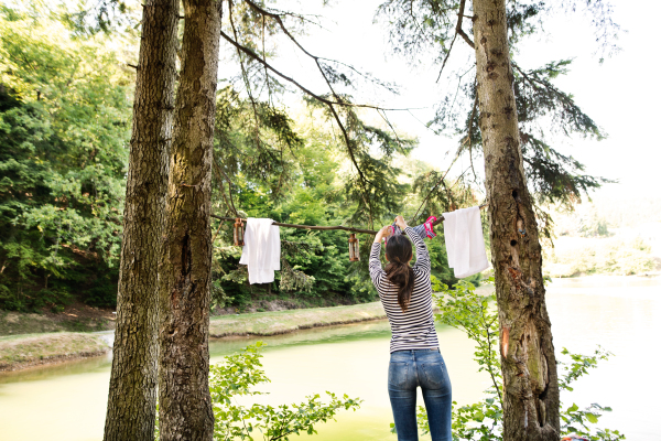 Beautiful woman on camping holiday in forest at the lake hanging clothes, rear view.