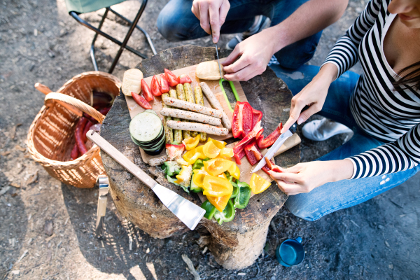 Unrecognizable couple enjoying camping holiday in forest, eating together. Barbecue with drinks and food.