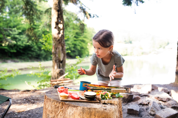 Little girl camping in forest with her family, eating grilled food. Barbecue with drinks and food.