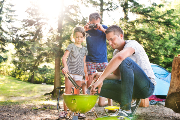 Beautiful family enjoying camping holiday in forest. Father cooking meat on barbecue grill, grandfather taking picture with camera.