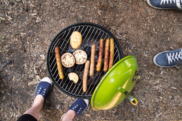 Beautiful family enjoying camping vacation, cooking sausages, cheese and vegetables on barbecue grill.