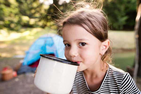 Face of cute little girl camping outdoors with her family, drinking water from a big enameled mug.