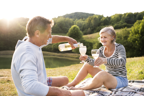 Beautiful senior couple at the lake having a picnic, sitting on blanket, drinking wine.