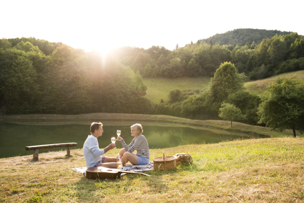 Beautiful senior couple at the lake having a picnic, sitting on blanket, drinking wine.