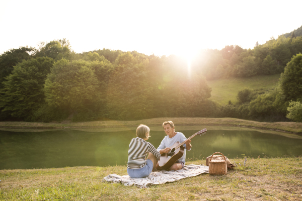 Beautiful senior couple at the lake having a picnic, sitting on blanket. Man playing guitar.