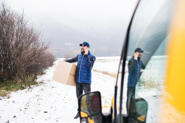 Delivery man delivering parcel box to recipient - courier service concept. A man with a smartphone making a phone call.