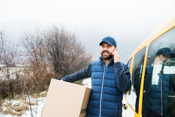 Delivery man delivering parcel box to recipient - courier service concept. A man with a smartphone making a phone call.