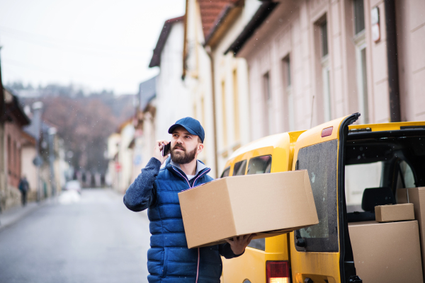 Delivery man holding a parcel box on the street - courier service concept. A man with a smartphone making a phone call.