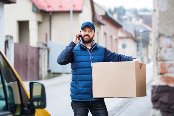 Delivery man holding a parcel box on the street - courier service concept. A man with a smartphone making a phone call.