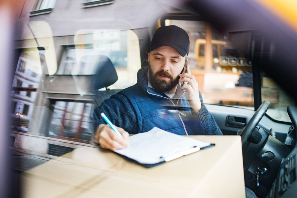 Delivery man with a parcel box in the car- courier service concept. A man with a smartphone making a phone call. Shot through glass.