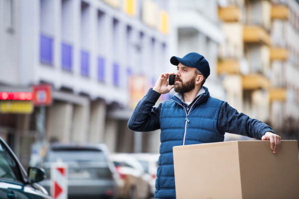 Delivery man holding a parcel box on the street - courier service concept. A man with a smartphone making a phone call.