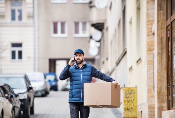 Delivery man holding a parcel box on the street - courier service concept. A man with a smartphone making a phone call.