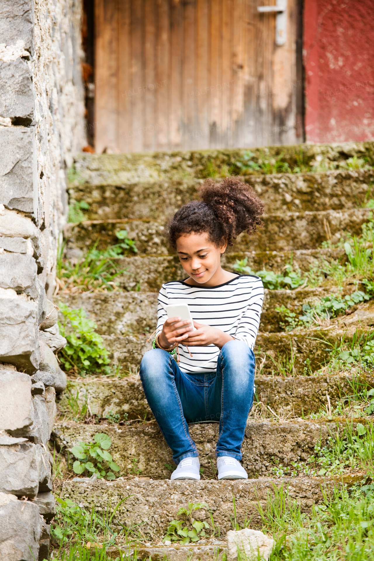 Beautiful african american girl with curly hair outdoors, sitting on stairs, holding smart phone, wearing headphones, listening music.