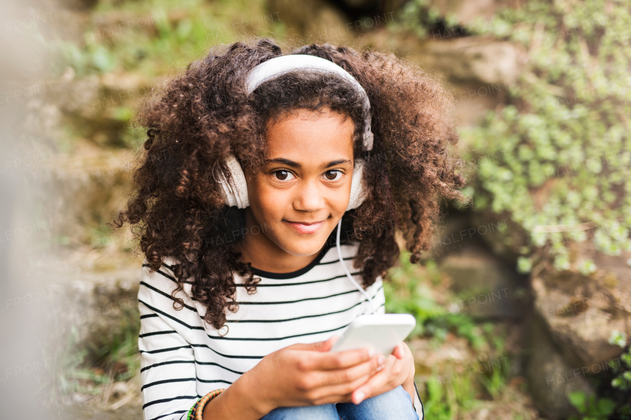 Beautiful african american girl with curly hair outdoors, sitting on stairs, holding smart phone, wearing headphones, listening music.