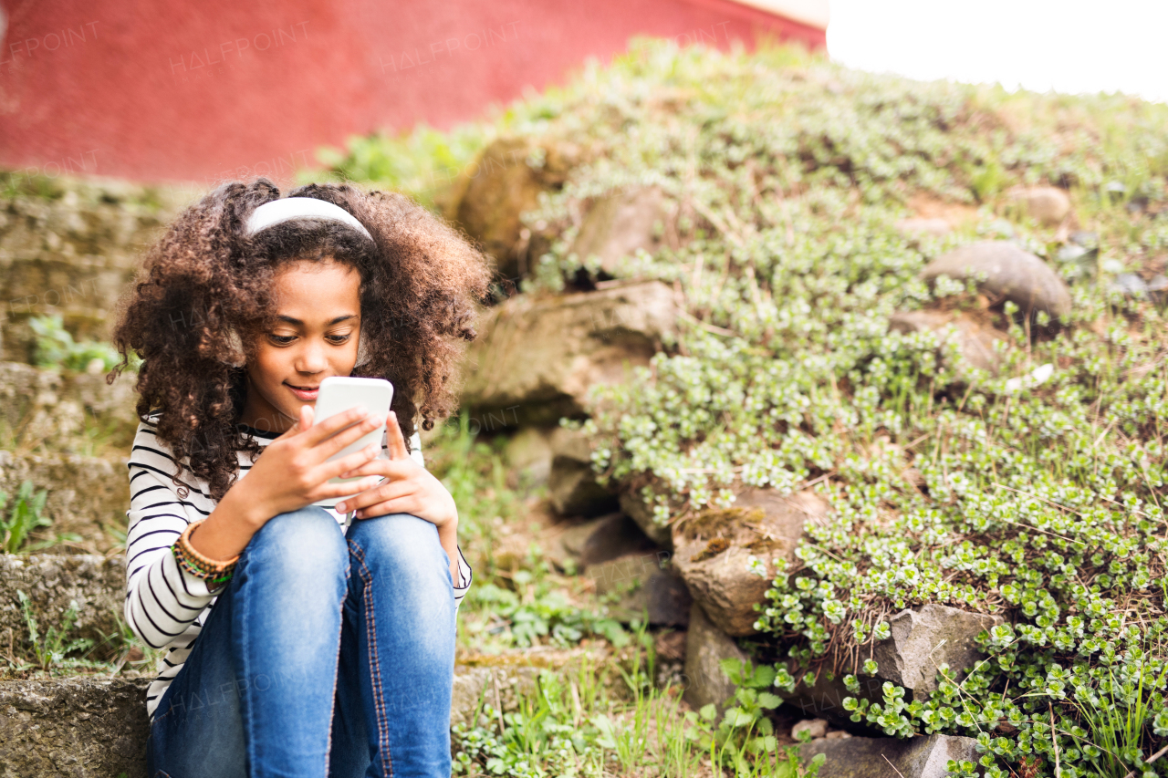 Beautiful african american girl with curly hair outdoors, sitting on stairs, holding smart phone, wearing headphones, listening music.