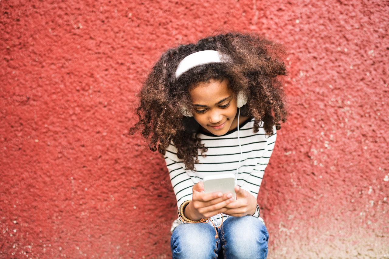 Beautiful african american girl with curly hair outdoors against red wall, holding smart phone, wearing yellow headphones, listening music.