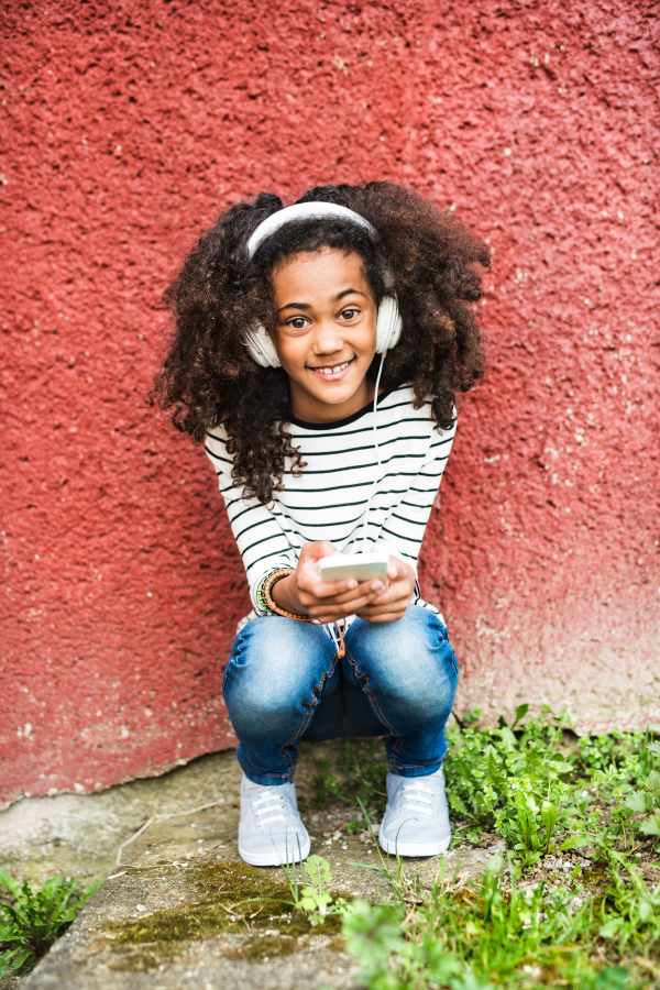 Beautiful african american girl with curly hair, holding smart phone, wearing headphones, listening music.