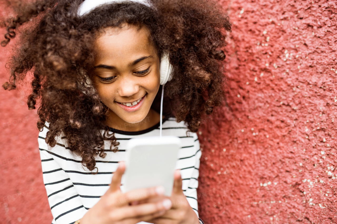 Beautiful african american girl with curly hair outdoors against red wall, holding smart phone, wearing yellow headphones, listening music.