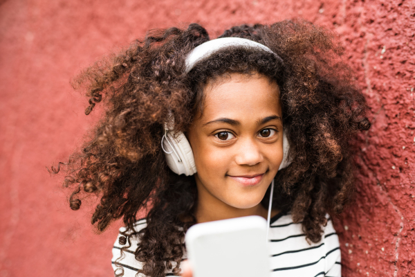 Beautiful african american girl with curly hair outdoors against red wall, holding smart phone, wearing yellow headphones, listening music.