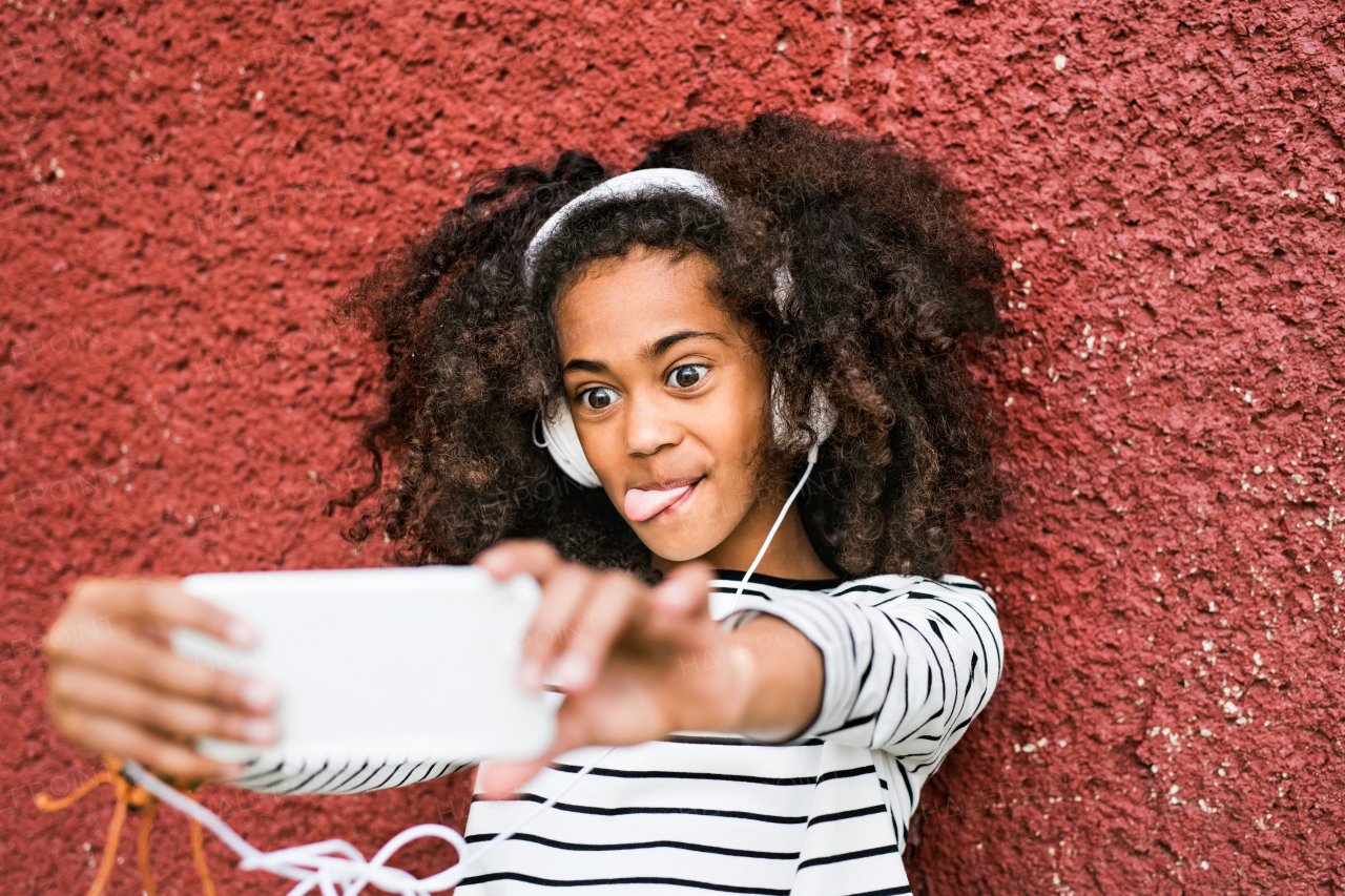 Beautiful african american girl with curly hair outdoors against red wall, wearing headphones, listening music, holding smart phone, taking selfie.