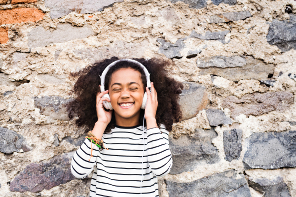 Beautiful african american girl with curly hair outdoors, against stone wall, wearing headphones, listening music.