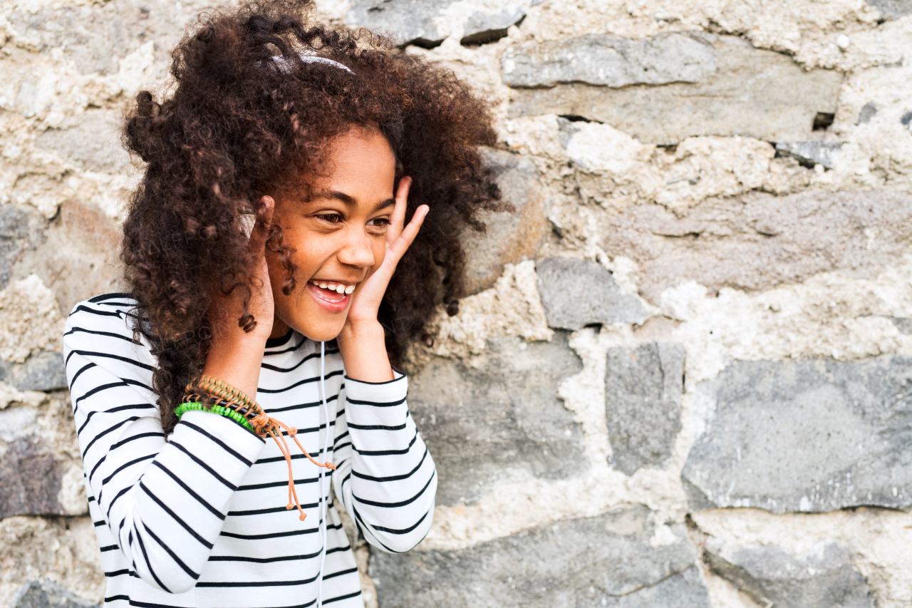 Beautiful african american girl with curly hair outdoors, against stone wall, wearing headphones, listening music.