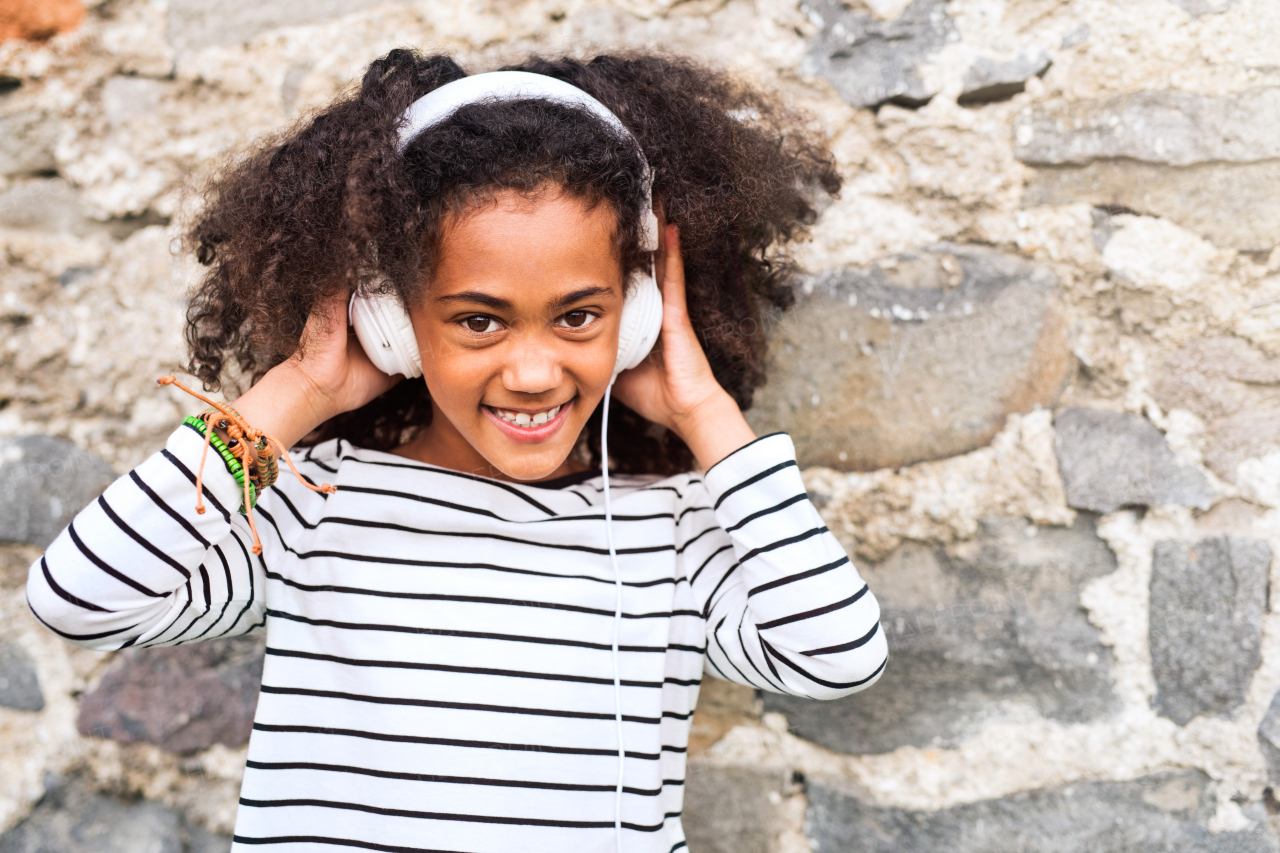 Beautiful african american girl with curly hair outdoors, against stone wall, wearing headphones, listening music.