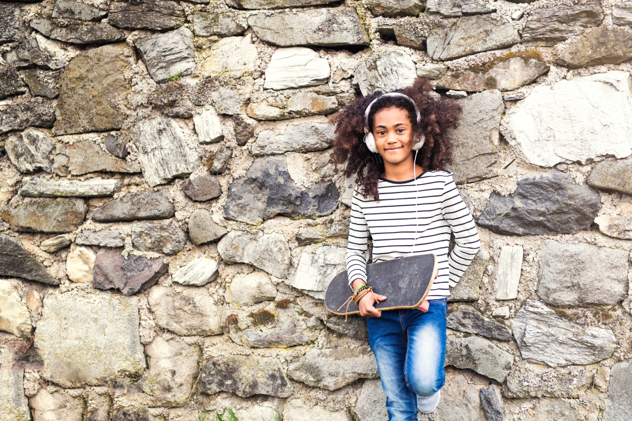 Beautiful african american girl with curly hair outdoors, against stone wall, holding skateboard, wearing headphones, listening music.