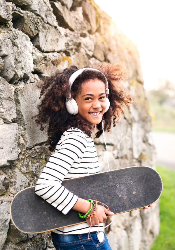 Beautiful african american girl with curly hair outdoors, against stone wall, holding skateboard, wearing headphones, listening music.