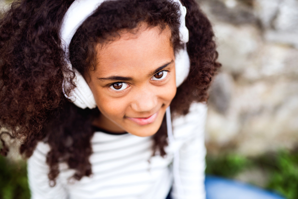 Beautiful african american girl with curly hair outdoors, against stone wall, wearing headphones, listening music.