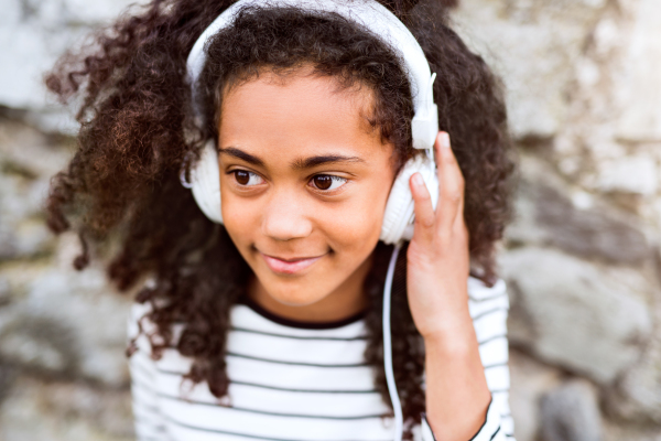 Beautiful african american girl with curly hair outdoors, against stone wall, wearing headphones, listening music.