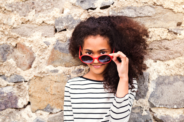 Beautiful african american girl with curly hair outdoors wearing striped t-shirt and red sunglasses, standing against stone wall.