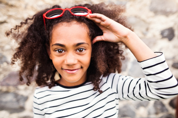 Beautiful african american girl with curly hair outdoors wearing striped t-shirt and red sunglasses, standing against stone wall.