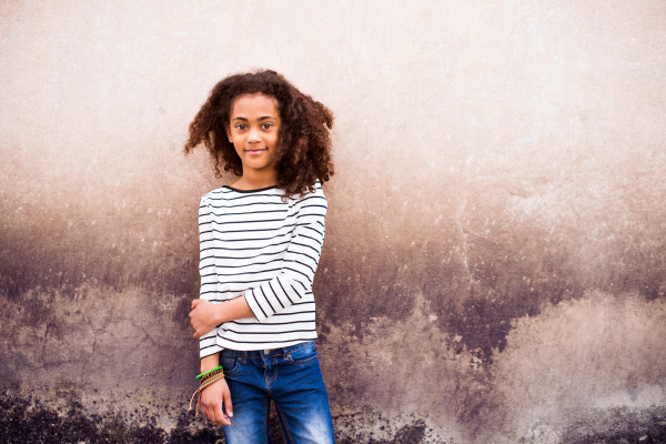 Beautiful african american girl with curly hair outdoors wearing striped t-shirt standing against old concrete wall.