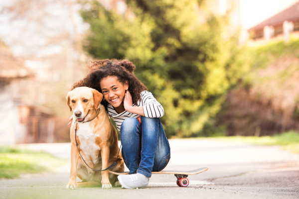 Beautiful african american girl with curly hair outdoors with her cute dog, sitting on skateboard.