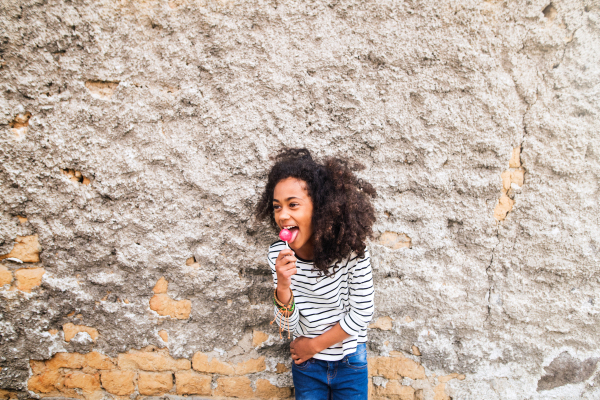 Beautiful african american girl with curly hair outdoors wearing striped t-shirt, eating lollipop, standing against old concrete wall.